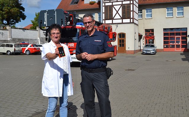 Bild: Elisabeth Ringleb, Pharmazie-Ingenieurin der Br&uuml;cken-Apotheke M&uuml;hlhausen, &uuml;bergab das Messger&auml;t an Hauptbrandmeister Martin Schmidt von der Feuerwehr M&uuml;hlhausen.