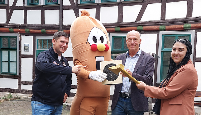 Markus Edom und Chiara Heller vom Kultur-Referat der Stadt M&uuml;hlhausen, Maskottchen Wurschti und der stellvertretende Gesch&auml;ftsf&uuml;hrer des Bratwurstmuseums, Kay Wipprecht, mit der Goldenen Axt, die bei den Wettbewerben zur Holzfahrt verliehen wird. (Foto: Stadtverwaltung M&uuml;hlhausen)